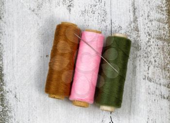 Three spools of different colors of thread, with single needle, on top of rustic white wood. 