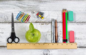 Green apple, pens, pencils, erasers, paper clips, tacks and scissors lined up with ruler on top of white desktop. Education concept. 