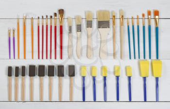 High angled view of brand new paint brushes and applicators organized on white wood. Layout in horizontal format.