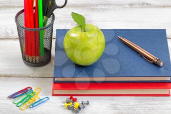A healthy green apple, books, pen, paper clips, tacks and container with supplies on top of white desktop. Education concept. 