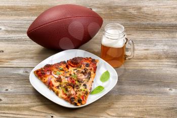 Two slices of freshly baked pizza in a white plate, football, and a pint of justly poured beer setting on rustic wooden table.
