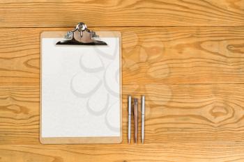 Top view of traditional clipboard, blank paper, and pens on wooden desktop. 