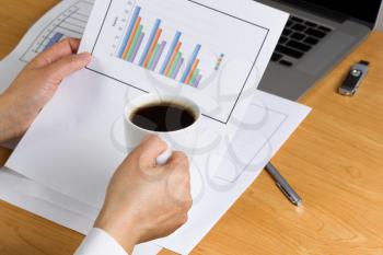 Woman hand holding a cup of dark coffee while looking over financial data with printed graphs, partial laptop, pen and thumb drive on desktop.