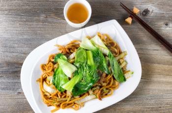 High angled view of spicy cooked noodles, onion, bok choy, chicken slices, chopsticks and tea on rustic wood. 