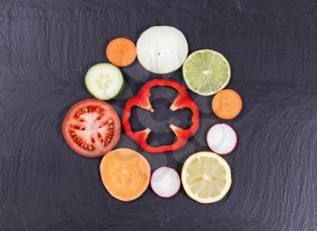 Overhead view of various sliced vegetables and fruits, in circle layout, on black slate. 