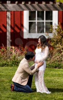 Husband kissing abdomen of expecting mom while kneeling in front of her on grass.