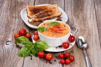 Front view of a bowl of fresh creamy tomato soup, tuna sandwich, with cherry tomatoes and basil leaves on rustic wooden boards. 