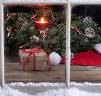 Gift box and Santa cap with evergreen decorations and burning candle in background through snowy window.