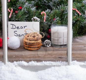 View of a stack of cookies, glass of milk and letter for Santa with evergreen decorations in background through snowy window.