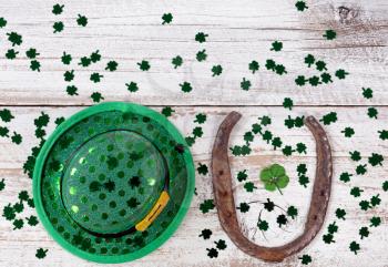 Real four leaf clover in the middle of rusty horseshoe with hat and shiny clovers on rustic wooden boards in overhead view  