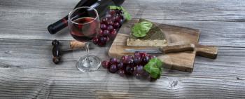 Glass of red wine with fresh cheese wedge plus basil leaves and grapes on rustic wooden table 