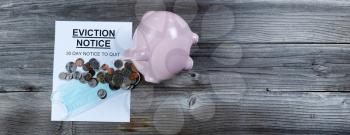 Eviction notice with personal facemask protection and tipped over piggy bank spilling coins on rustic wooden table in flat lay format