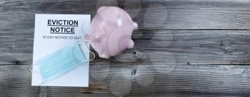 Eviction notice with personal facemask protection and tipped over piggy bank on rustic wooden table in flat lay format 