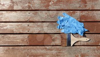 Brush loaded with stain on outdoor wooden cedar plus latex gloves for resurfacing the home deck in flat lay format 