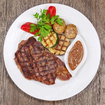 Grilled steaks, baked potatoes and vegetables on white plate on wooden background.