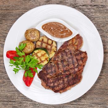 Grilled steaks, baked potatoes and vegetables on white plate on wooden background.