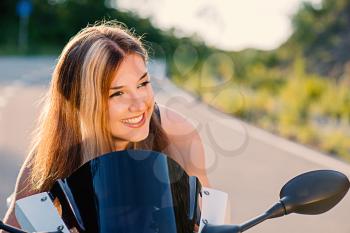 Young beautiful blonde girl in black t-shirt is resting on nature in seat of modern motorcycle. Outdoor portrait in soft sunny tones.