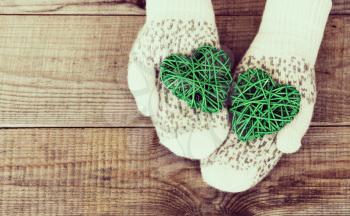 Woman hands in light teal knitted mittens are holding green hearts on wooden background. Winter and Christmas concept.