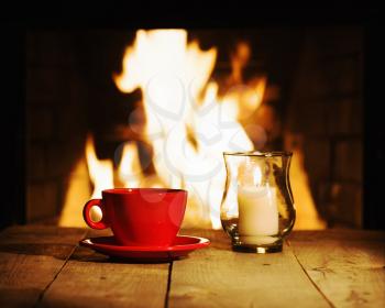 Red cup of coffee or tea and candle on wooden table near fireplace. Winter and Christmas holiday concept.