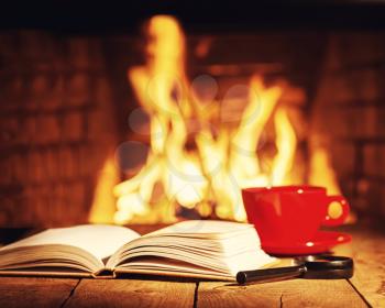 Red cup of tea or coffee, magnifier glass and old books near fireplace on wooden table. Winter and Christmas holiday concept. Photo with retro filter effect.