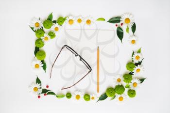 Paper, pencil and glasses with wreath frame from chamomile and chrysanthemum flowers, ficus leaves and ripe rowan on white background. Overhead view. Flat lay.
