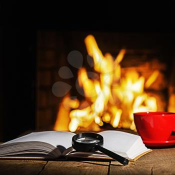 Red cup of coffee or tea, magnifier glass and old book on wooden table near  fireplace. Winter and Christmas holiday concept.