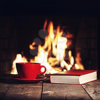 Red cup of coffee or tea and old book on wooden table near  fireplace. Winter and Christmas holiday concept. Photo with retro filter effect.