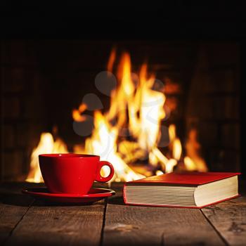 Red cup of coffee or tea and old book on wooden table near  fireplace. Winter and Christmas holiday concept.