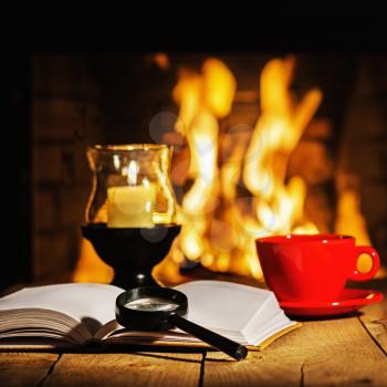 Red cup of coffee or tea, candle in lamp, magnifier glass and old book on wooden table near fireplace. Winter and Christmas holiday concept.