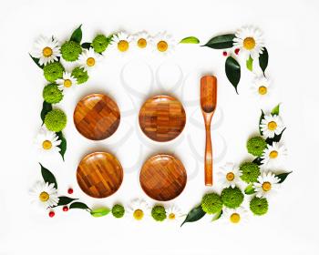 Wooden dishes with wreath frame from chamomile and chrysanthemum flowers, ficus leaves and ripe rowan on white background. Overhead view. Flat lay.