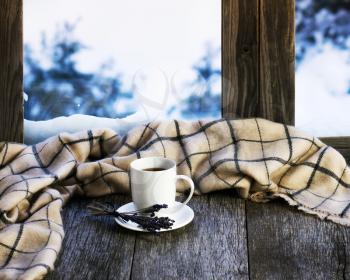White cup of coffee or tea, lavender flowers and woolen plaid located on stylized wooden window sill. Winter concept of comfort and relaxation.