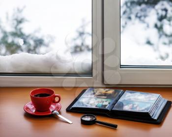 Red cup of coffee or tea with a metal spoon, photo album and loupe located on a stylized wooden windowsill. Winter concept of comfort and relaxation.