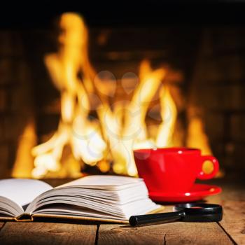 Red cup of tea or coffee, magnifier glass and old books near fireplace on wooden table. Winter and Christmas holiday concept.