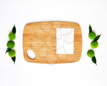 Wooden cutting board and paper with decoration of chrysanthemum flowers and ficus leaves on white background. Overhead view. Flat lay.