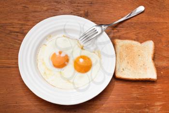 breakfast with two fried eggs on white plate, fresh toast on wooden table
