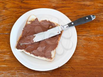 top view of sweet sandwich from fresh toast with chocolate spread on white plate, table knife on wooden table