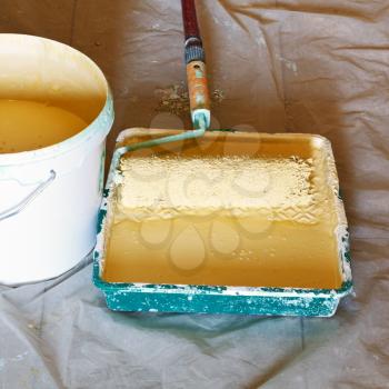 painter roller brush in plastic paint tray with yellow emulsion paint and bucket with paint