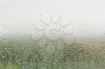 raindrops on home window glass with green forest and grey sky background