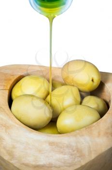 olive oil in thin trickle flows from bottle on green olives in wooden bowl close up isolated on white background