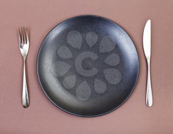 top view of empty black plate with fork and knife set on brown background