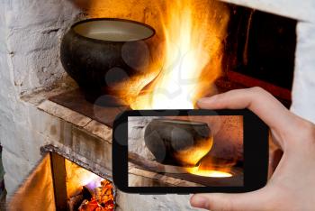 photographing food concept - tourist takes picture of fire hearth of russian stove and old cast iron pot with oven fork on smartphone,