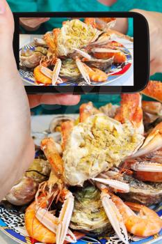 photographing food concept - tourist takes picture of plate with cut crab, oysters, shrimps and other seafood on smartphone, France