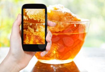 photographing food concept - tourist takes picture of fresh honey in comb in glass bowl at table on smartphone, Russia