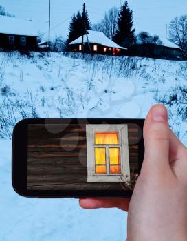 travel concept - tourist takes picture of yellow lighting window of rural log house in winter on smartphone,