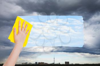 weather concept - hand deletes rainy clouds over city by yellow cloth from image and blue sky with white clouds are appearing