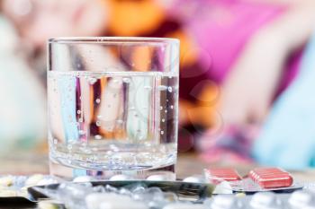 glass with water and pile of pills on table close up and in bed-sitting room on background