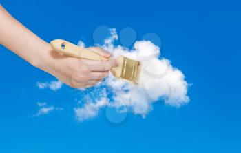 weather concept - hand with paintbrush paints white cloud in blue summer sky