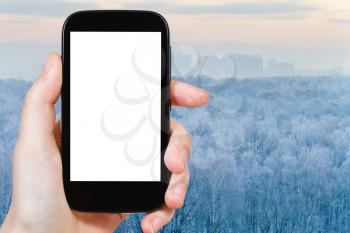 travel concept - hand holds smartphone with cut out screen and frozen winter forest on background