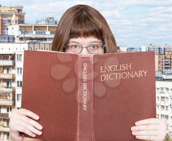 girl with spectacles looks over English Dictionary book with cityscape on background