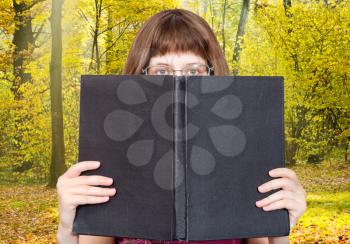 girl with glasses looks over big book with blank cover and sunny autumn forest on background